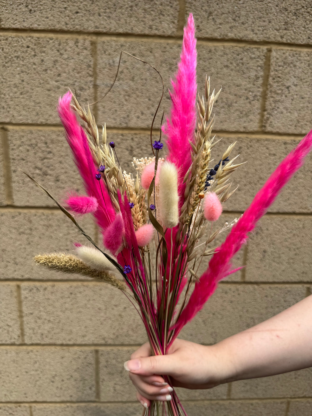 A hand holding a dried flower bouquet featuring vibrant pink pampas grass, beige wheat stalks, cream bunny tail grass, and small purple wildflower accents, set against a beige brick wall background.
