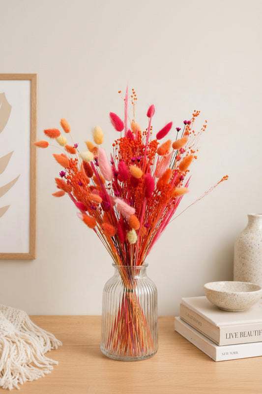 A vibrant dried flower arrangement in a ribbed clear glass vase, styled on a wooden table with books, a ceramic bowl, and a neutral wall background.