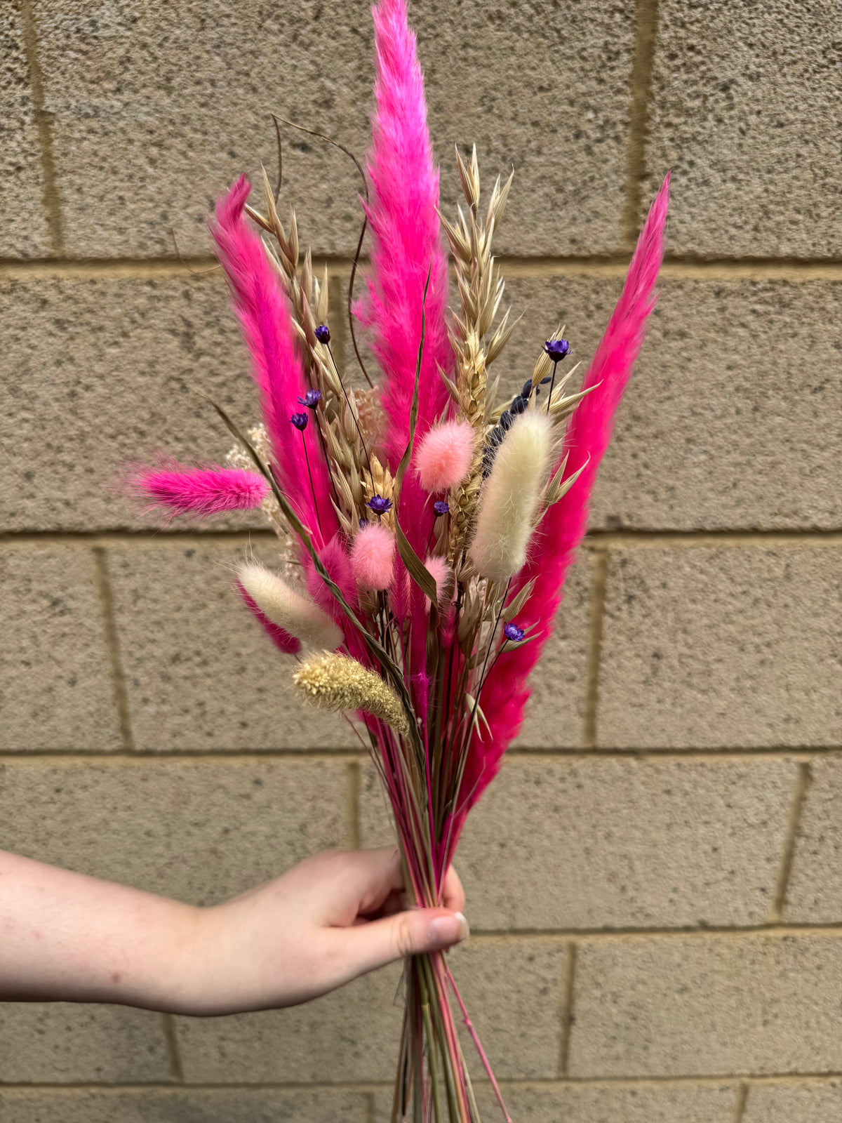 A hand holding a dried flower bouquet featuring vibrant pink pampas grass, beige wheat stalks, cream bunny tail grass