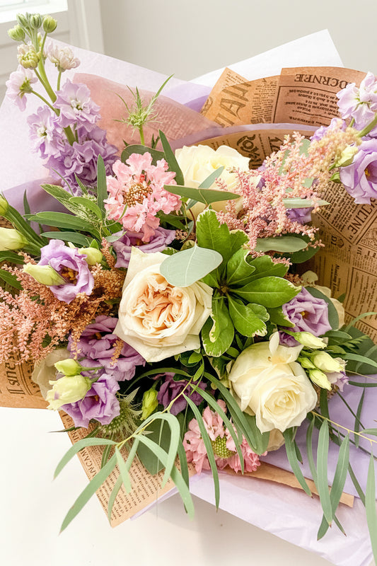 A close-up of a floral bouquet with cream roses, pink flowers, purple blossoms, green leaves, and eucalyptus stems, arranged in a box lined with brown newspaper-style wrapping paper and pale lavender tissue paper.
