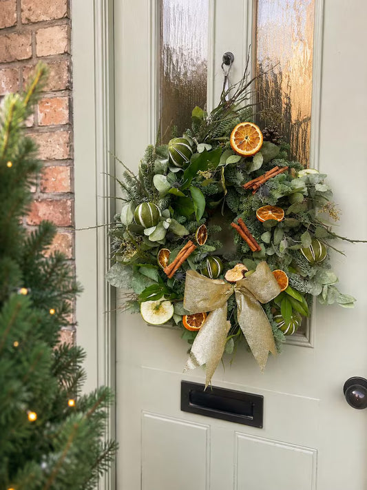 A festive Christmas wreath hanging on a light sage green front door, decorated with green pine branches, frosted eucalyptus, striped green ornaments, dried orange slices, cinnamon sticks, and a large beige glitter bow.