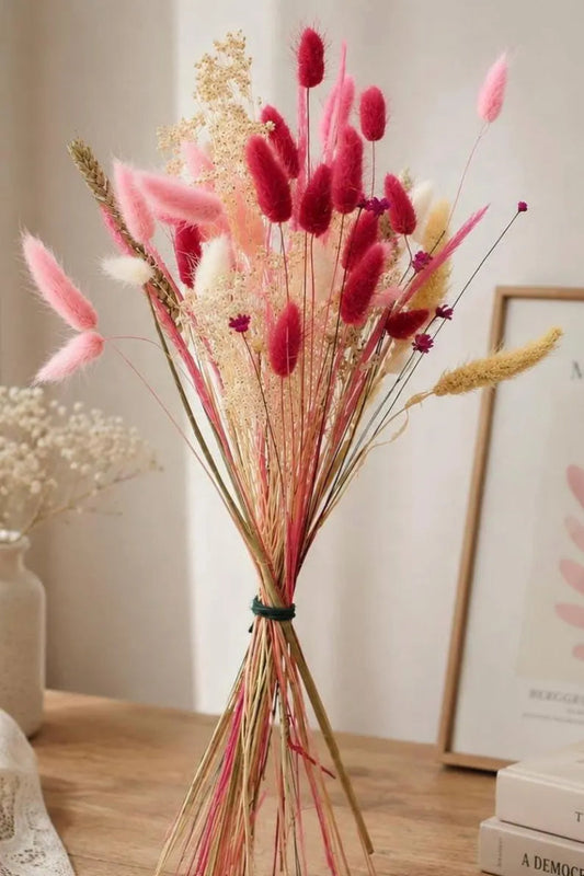 A dried flower arrangement featuring pink bunny tails, fluffy pink grass, and beige wild grasses tied with a dark green band, displayed in a vase on a wooden table.